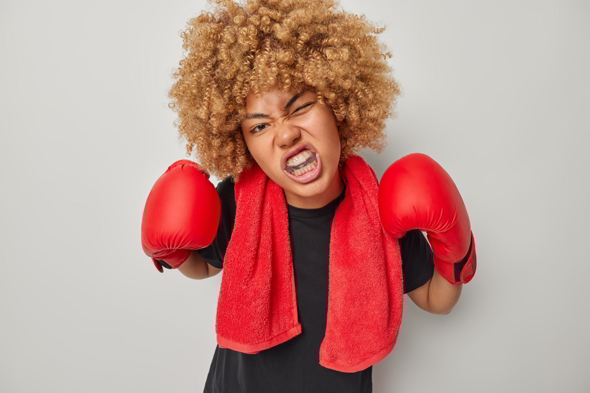 Indoor shot of curly haired female boxer protects teeth with mouthguard ready for fight wears black