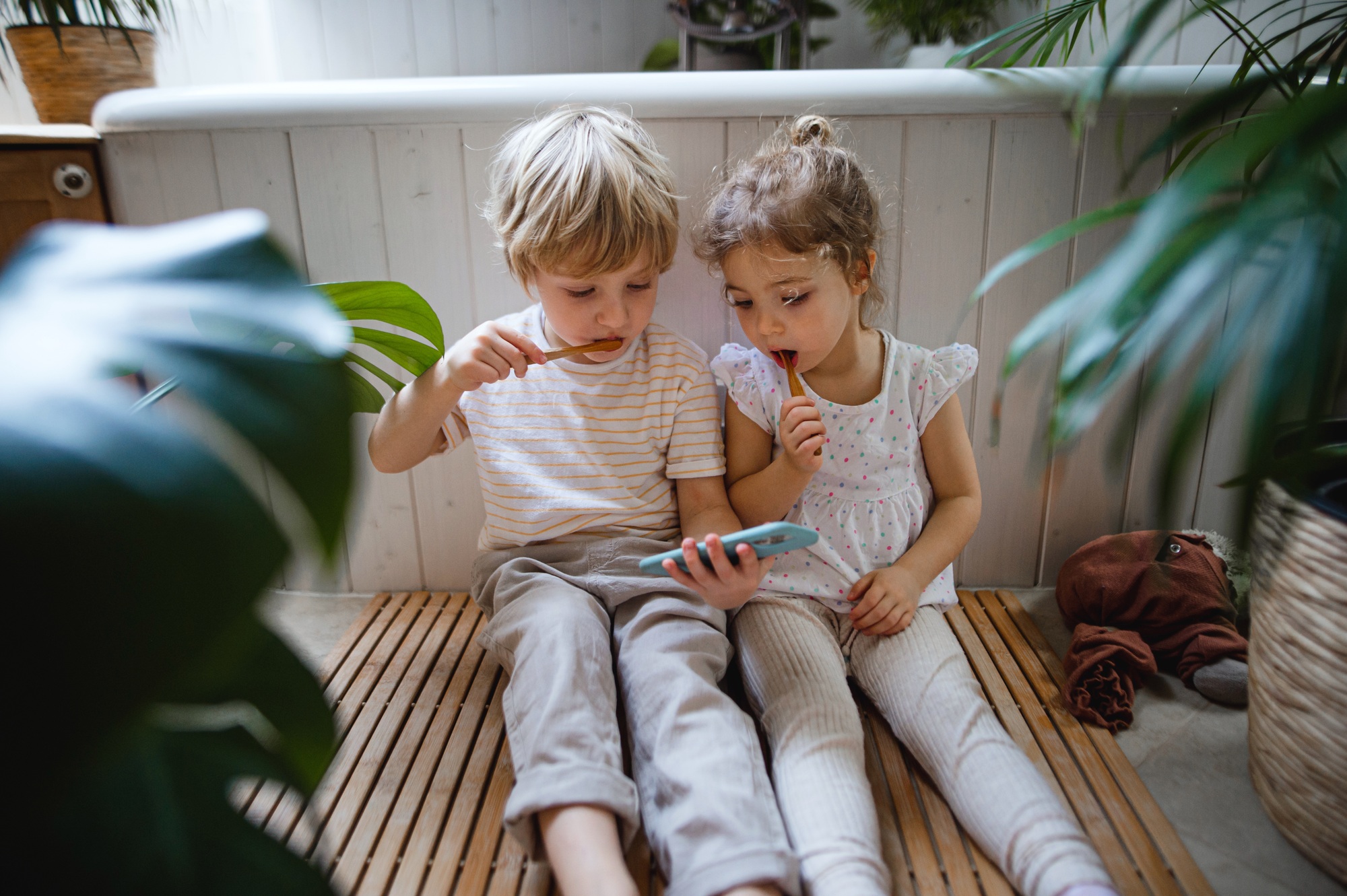 Cute small children sitting on floor indoors at bathroom, brushing teeth and using smartphone