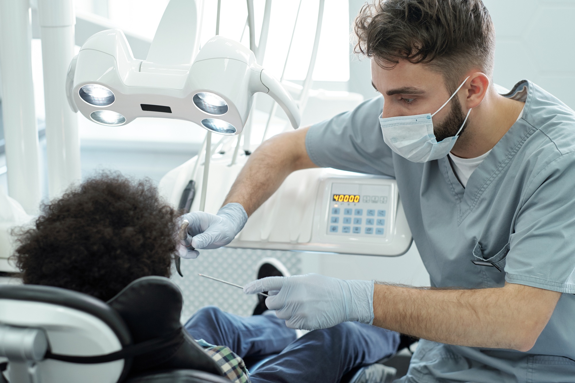 Dentist in uniform, mask and gloves holding dental drill and instrument by mouth of little patient