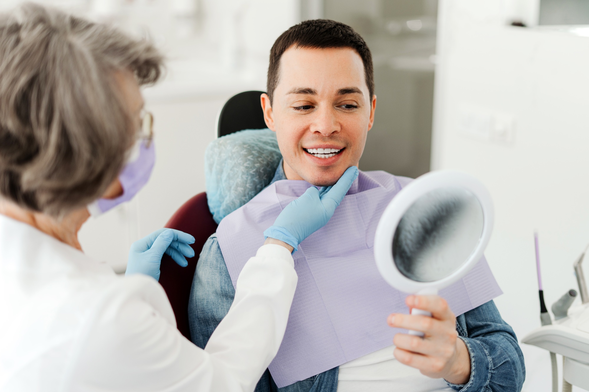 Smiling attractive male patient holding mirror, looking, talking to senior professional dentist
